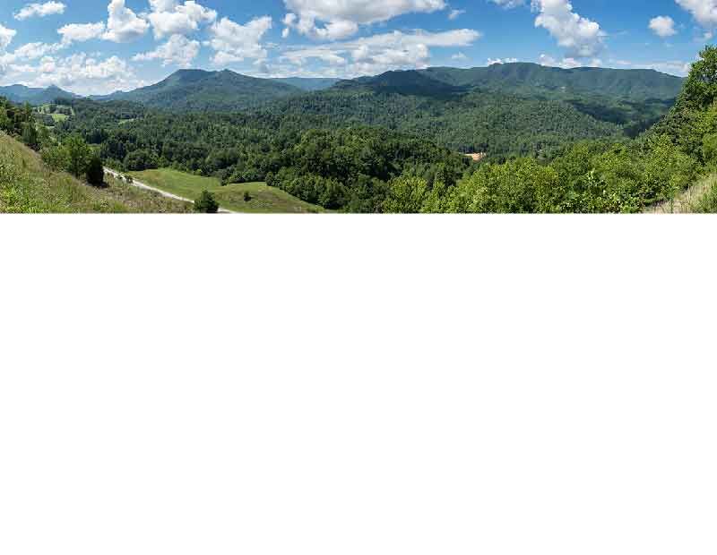 Weite Aussicht über eine grüne Hügel- und Berglandschaft an einem sonnigen Tag mit blauem Himmel und weißen Wolken