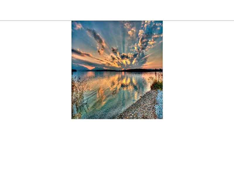 Spiegelung eines spektakulären Sonnenuntergangs im ruhigen Wasser eines Sees mit Kieselstrand und Vegetation am Ufer