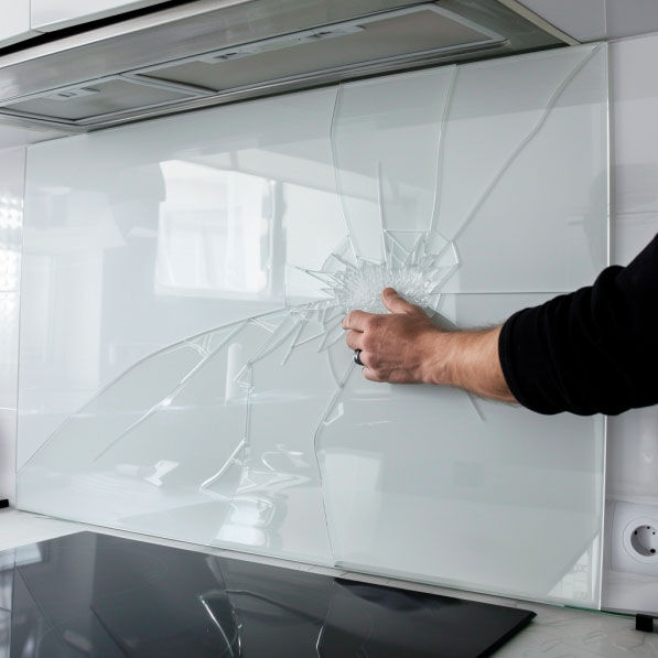 Beschädigte Glaswand in moderner Küche Nahaufnahme einer gebrochenen Glaswand in einer modernen Küche, berührt von einer Hand. Die Risse breiten sich sternförmig aus und sind deutlich sichtbar.