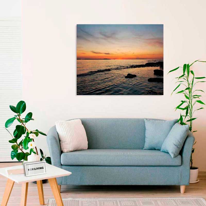 Ein gemütlich eingerichtetes Wohnzimmer mit einem blauen Sofa, zwei Kissen, einem kleinen Tisch mit Pflanzen und einem großen Bild eines Sonnenuntergangs an der Wand.