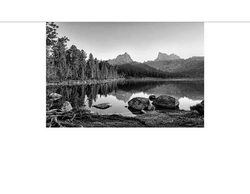 Schwarzweiß-Fotografie eines ruhigen Bergsees mit Felsen am Ufer, umgeben von dichten Wäldern und Berggipfeln im Hintergrund.