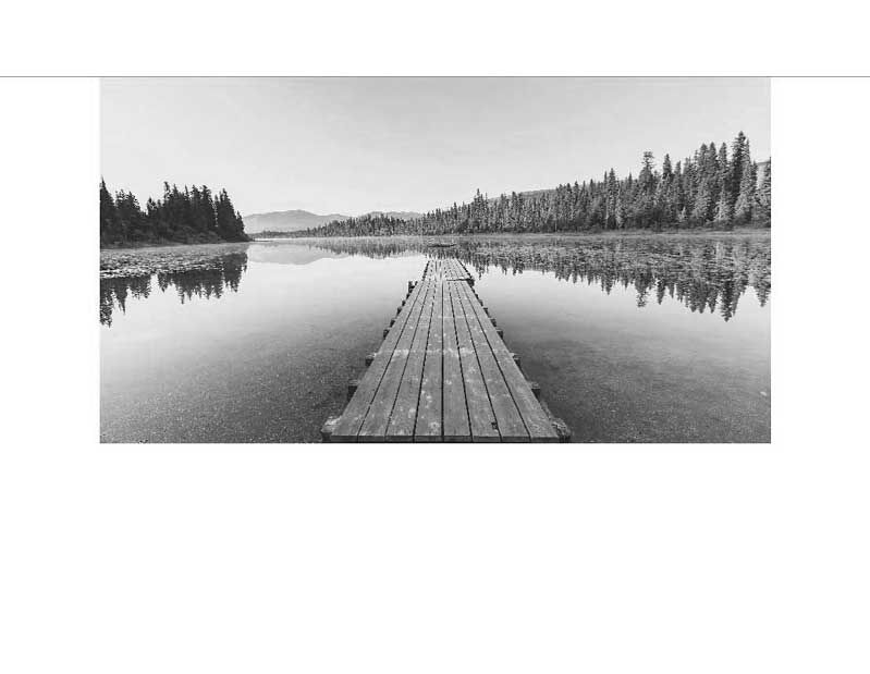 Ein Holzsteg führt über die ruhige Wasseroberfläche eines Sees, umgeben von Wäldern, in einer Schwarz-Weiß-Fotografie.