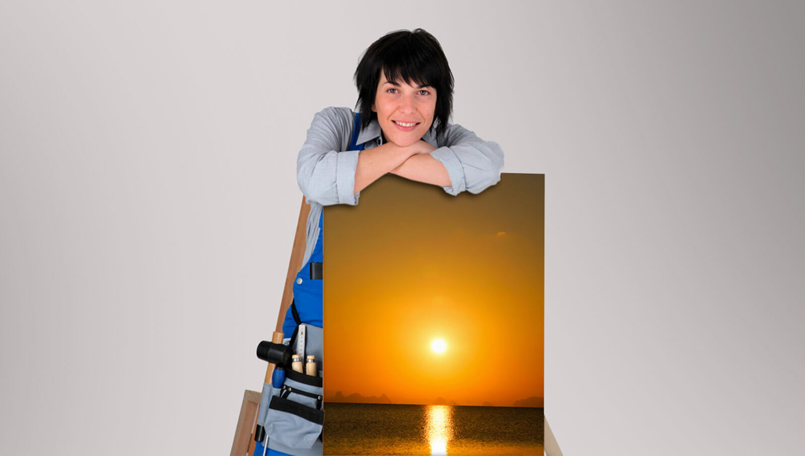 Frau auf Leiter mit Sonnenuntergangsfoto vor hellem Hintergrund Eine Frau in Arbeitskleidung lehnt sich auf eine Leiter und hält ein Bild des Sonnenuntergangs über dem Meer, vor einem hellen Hintergrund.