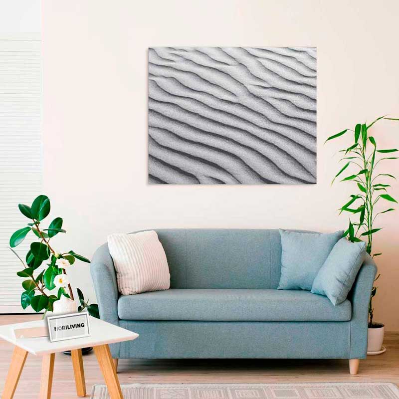 Ein gemütliches Wohnzimmer mit einer grauen Couch, Kissen und einem Wandbild mit Sandmuster. Daneben stehen Pflanzendekorationen.