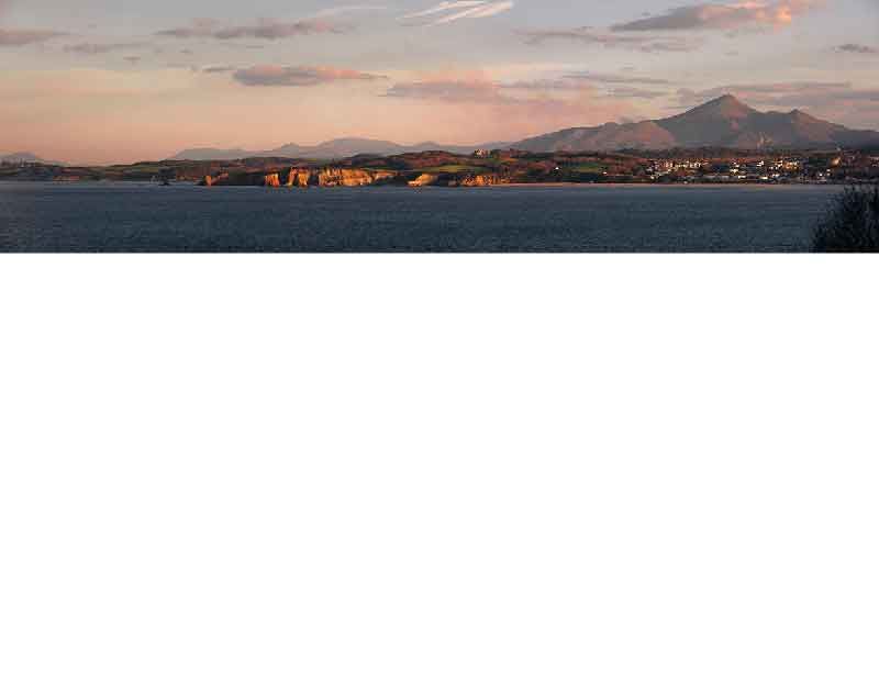 Weitläufiger Blick auf eine Küstenlandschaft mit Bergen im Hintergrund und einem ruhigen Meer bei Sonnenuntergang