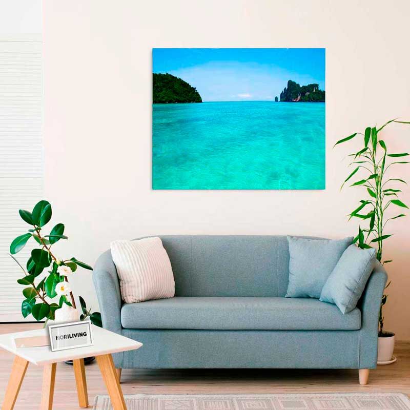 Ein stilvoll eingerichtetes Wohnzimmer mit einem hellblauen Sofa, darauf zwei Kissen, einem Couchtisch mit Pflanze und einem großen Wandbild einer klaren, blauen Küste.