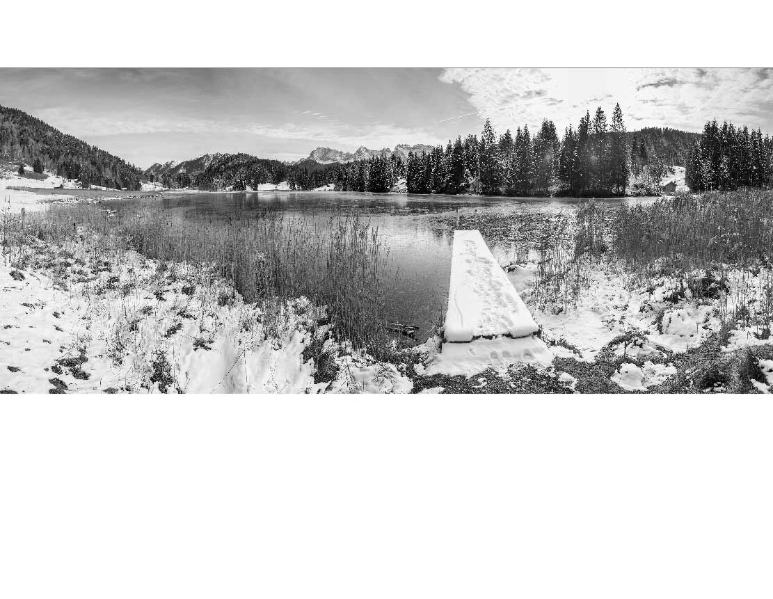 Panorama einer verschneiten Landschaft mit einem See und einem schneebedeckten Steg im Vordergrund, umgeben von Bäumen und Bergen.