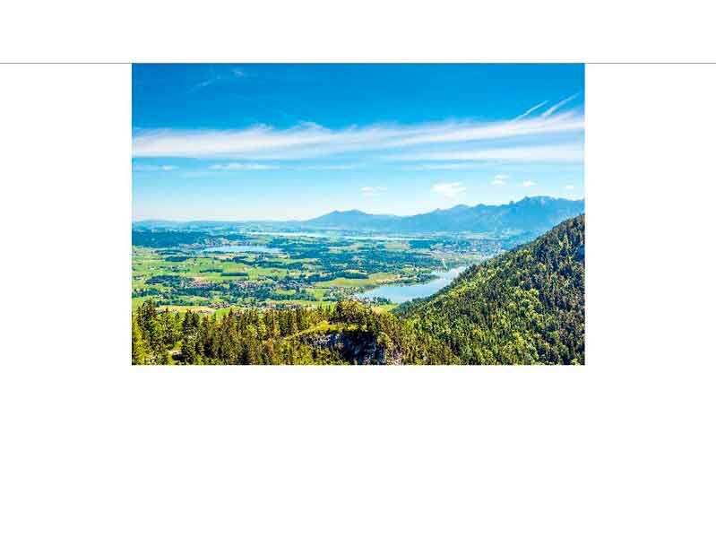 Weitläufige Aussicht auf grüne Felder, einen klaren See und die Alpen in Bayern unter klarem Himmel