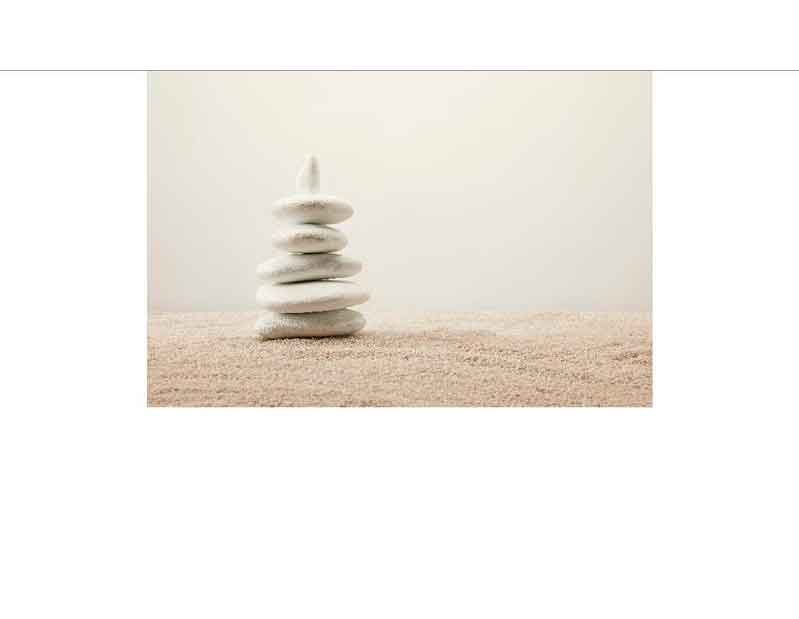 Ein Zen-Steinturm aus fünf weißen, glatt geschliffenen Steinen auf einem sandigen Untergrund vor einem neutralen Hintergrund.