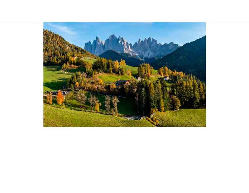 Panoramablick auf eine herbstliche Landschaft mit grünen Wiesen, bunten Bäumen und Berggipfeln der Dolomiten im Hintergrund